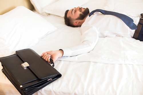 Portrait of tired bearded businessman laying on hotel bed relaxing after business trip focus on leather case in foreground