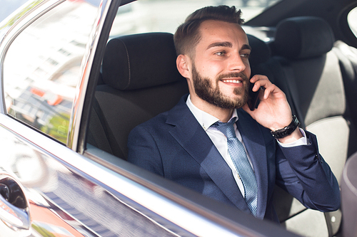 Portrait of handsome bearded businessman speaking by phone sitting in back seat of car and smiling happily