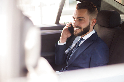 Portrait of handsome bearded businessman speaking by phone sitting in back seat of luxury car and smiling