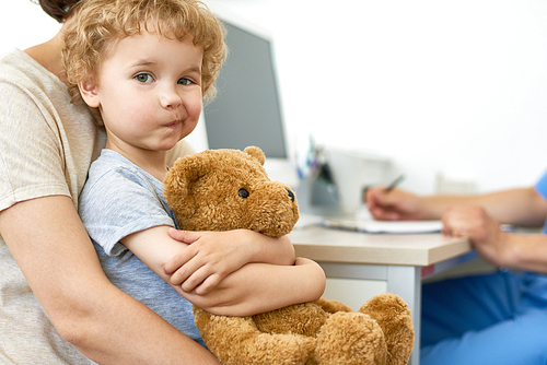 Portrait of cute child sitting on mothers lap in doctors office hugging plush teddy bear  and , copy space