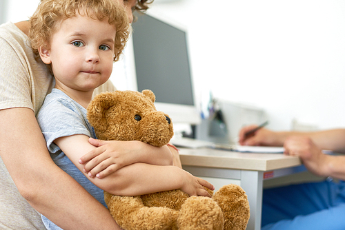 Closeup portrait of cute child sitting on mothers lap in doctors office waiting for check up hugging plush teddy bear toy