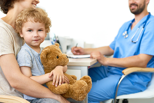 Portrait of cute child sitting on mothers lap in doctors office hugging plush teddy bear  and smiling at camera, copy space