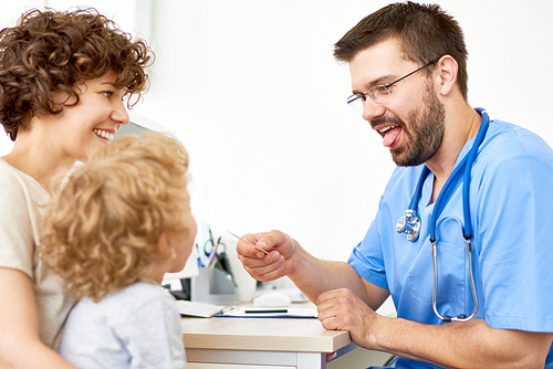 Portrait of friendly pediatrician examining little child sitting on mothers arms during appointment in office