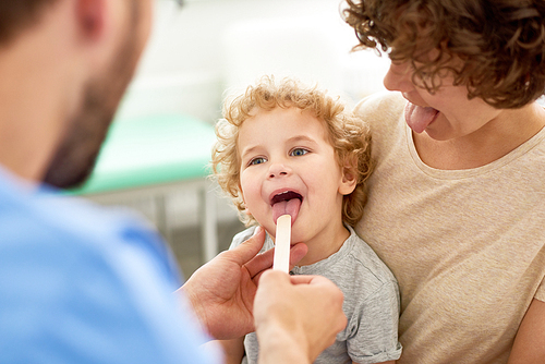 Portrait of cute curly child sitting on mothers lap during medical checkup, with doctor inspecting his open mouth
