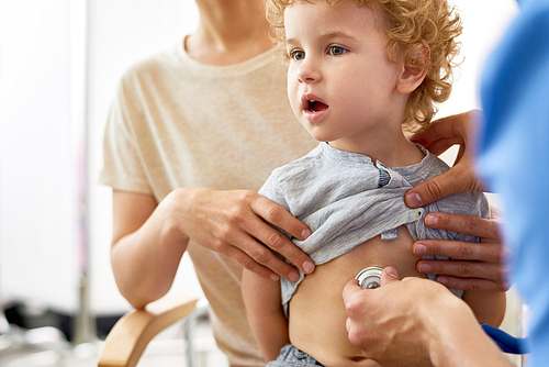 Portrait of cute curly boy sitting on mothers lap during medical checkup, unrecognizable doctor listening to childs heartbeat with stethoscope
