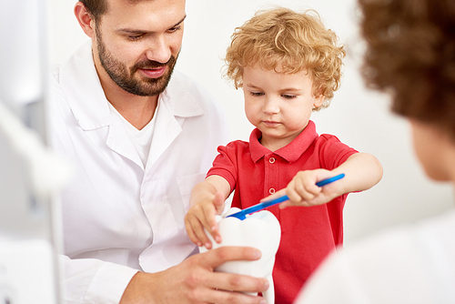 Portrait of handsome dentist teaching adorable curly boy how to brush teeth, using big tooth model