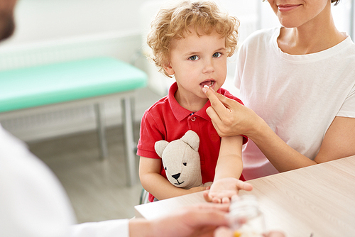 Portrait of cute curly-haired little boy taking pills in doctors office and  holding teddy bear toy
