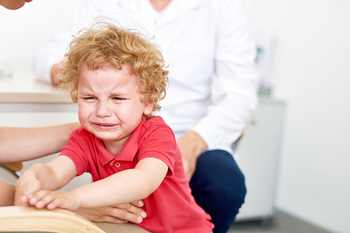 Portrait of curly little patient crying hysterically at pediatrician office refusing to be examined, blurred background