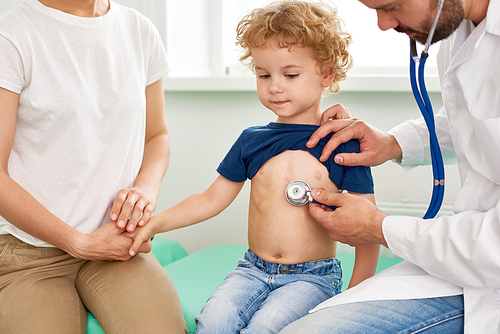 Portrait of adorable little boy visiting doctor, looking brave and holding mothers hand while pediatrician listening to heartbeat with stethoscope