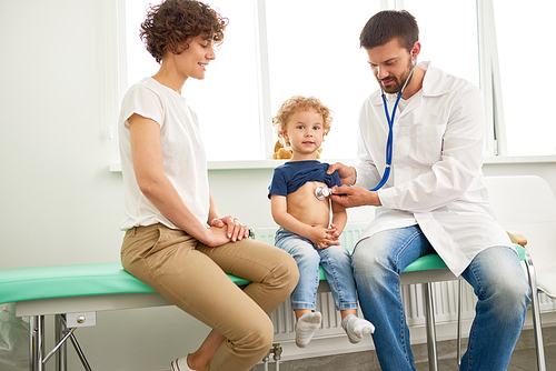 Portrait of adorable little boy visiting doctor, looking brave and smiling, while pediatrician listening to heartbeat with stethoscope
