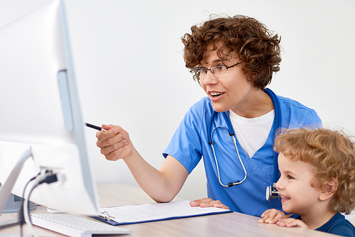 Portrait of friendly female doctor sitting at desk in office , showing something at screen to little boy