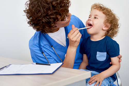 Portrait of cute little boy opening mouth for checkup sitting in doctors lap against white background
