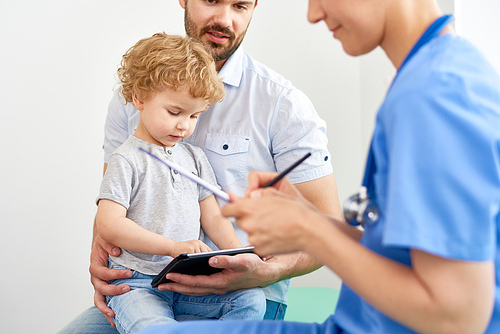 Portrait of little boy playing games at digital tablet while visiting doctor with dad