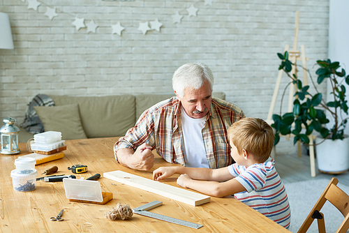 Portrait of grandfather and little boy talking while making wooden model at desk in small studio, old man teaching grandson woodwork