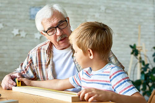 Portrait of grandfather and little boy talking while making wooden model at desk in small studio