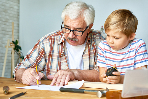 Portrait of old man and little boy making wooden models together sitting at work desk, grandfather drawing sketches