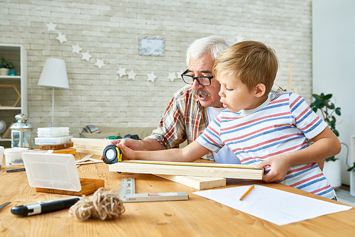 Side view portrait of old man teaching cute little boy woodwork, making wooden models together working at desk in small studio