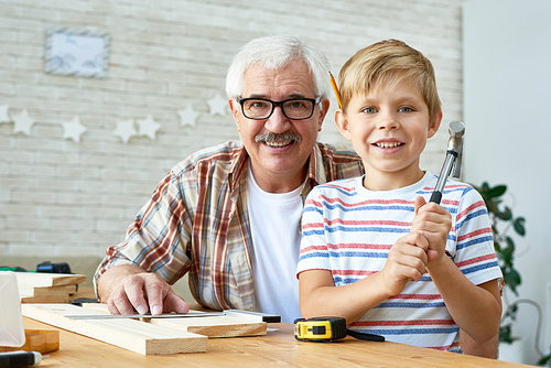 Portrait of happy senior man posing with little  grandson  and smiling while working with wood together at home