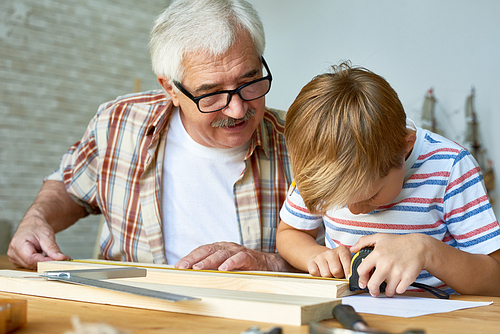 Portrait of old man teaching cute little boy woodwork, making wooden models together working at desk in small studio