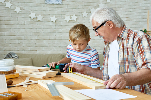 Portrait of white-haired senior man teaching little grandson carpentry, working with wood together sitting at desk