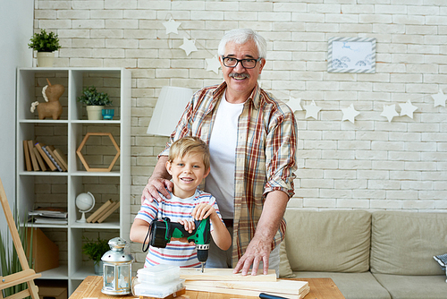 Portrait of happy grandfather posing with grandson  and smiling while working with wood  at home