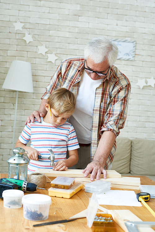 Portrait of senior man helping little boy hammering nails while making wooden model together at home