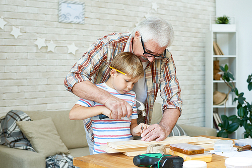 Portrait of grandfather teaching little boy woodwork, helping him build birdhouse at table in studio