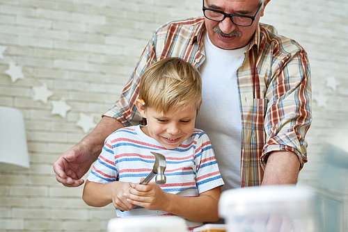 Portrait of smiling senior man helping little boy hammering nails while making wooden model together at home