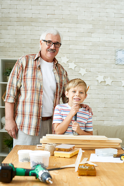 Portrait of happy grandfather posing with grandson  and smiling while working with wood together at home