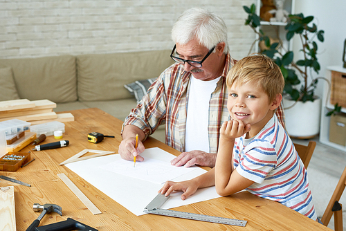 Portrait of cute little boy  smiling happily while making wooden models with grandpa, sitting at desk in small studio, copy space