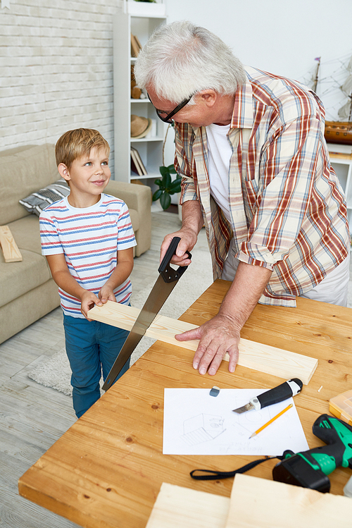 High angle portrait of nice grandfather teaching grandson woodwork, helping little boy saw piece of wood while making wooden models together