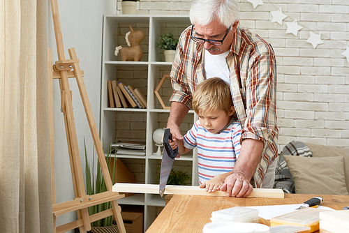 Portrait of nice grandfather teaching grandson woodwork, helping little boy saw piece of wood while making wooden models together