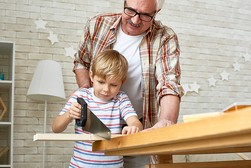 Portrait of nice grandfather teaching grandson woodwork, helping little boy saw piece of wood while making wooden models together