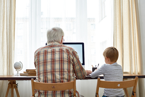 Rear view portrait of grandfather and grandson doing homework together sitting at desk with  blank screen computer