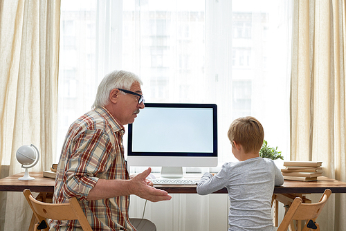 Portrait of little boy and his grandpa sitting at desk with modern computer together doing homework, senior man explaining something gesturing actively