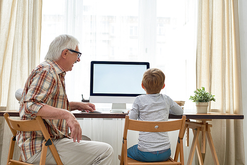 Rear view portrait of grandfather and grandson doing homework together sitting at desk with modern blank screen computer