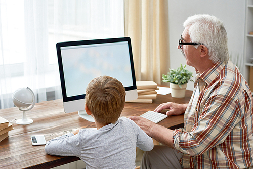 Portrait of grandfather helping little boy do homework sitting at desk with blank screen of modern computer
