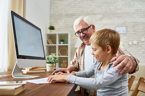 Side view portrait of grandfather helping little boy do homework both smiling happily sitting at desk with modern computer