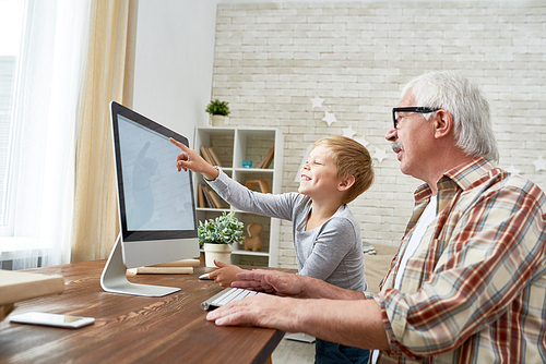 Portrait of grandfather helping happy little boy do homework sitting at desk and pointing to blank screen of modern computer