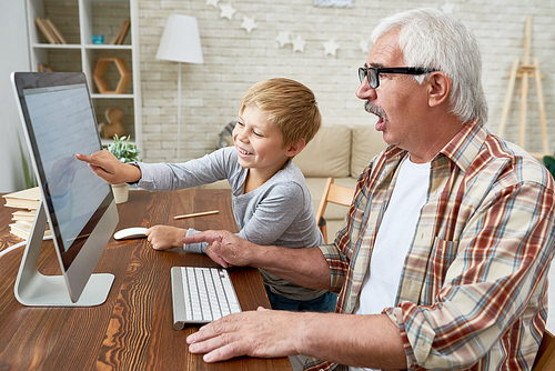 Portrait of smiling little boy teaching old grandpa how to use modern computer sitting  at desk together, old man looking at PC with  mouth open in amazement