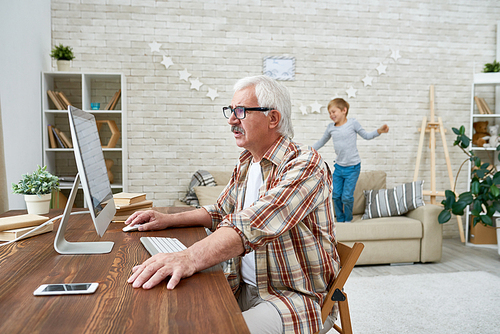 Portrait of senior man learning to use modern computer sitting at desk with little boy jumping on sofa in background