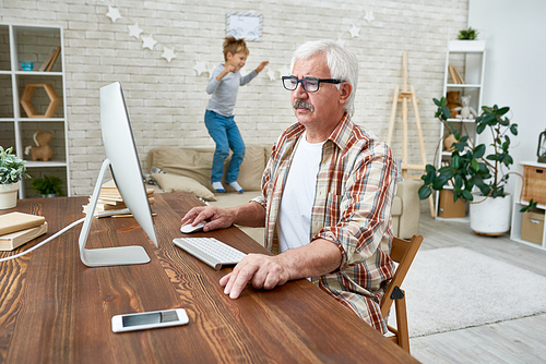 Portrait of white haired senior man learning to use modern computer sitting at desk with little boy jumping on sofa in background