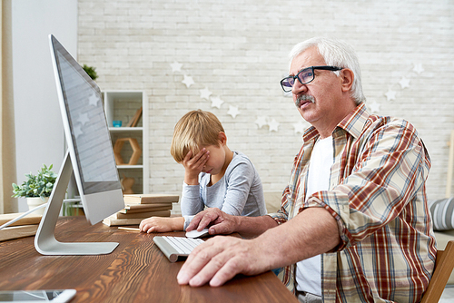 Portrait of senior man learning to use internet with tired little boy trying to teach him, both sitting at desk at home