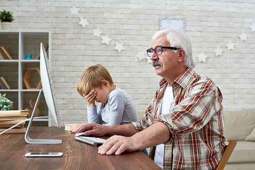 Portrait of  little boy trying to teach old grandpa how to use modern computer sitting  at desk together, old man looking at PC with  amazement