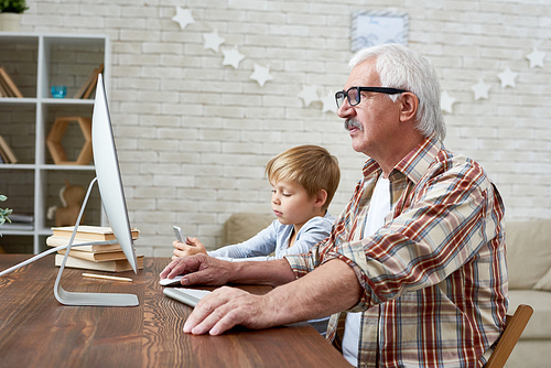 Portrait of senior man learning to use internet with tired little boy sitting next to him at desk at home