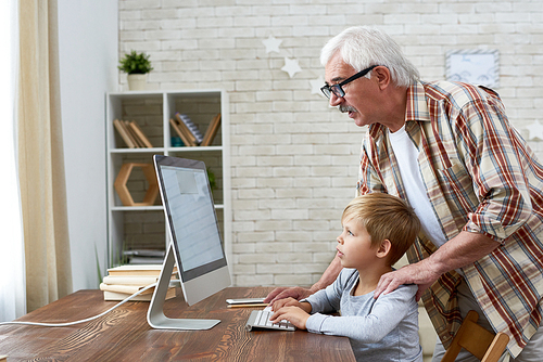 Portrait of grandfather helping little boy do homework sitting at desk looking at blank screen of modern computer