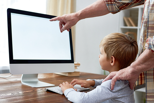 Portrait of little boy learning to use modern pc sitting at desk and doing homework with grandpa explaining him something pointing at blank white screen