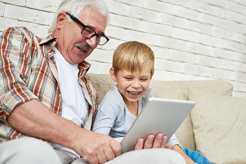 Portrait of happy senior man using digital tablet watching videos with grandson both laughing sitting on couch at home