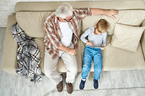 Top view portrait of little boy and his grandpa spending time together relaxing on cozy sofa at home, boy using digital tablet and showing something