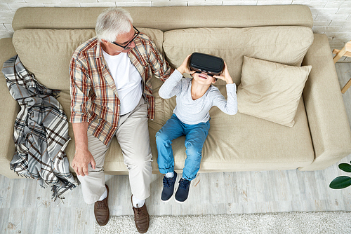 Top view portrait of little boy and his grandpa spending time together relaxing on cozy sofa at home, boy using VR glasses watching 360 videos